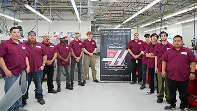 Group of 11 men in maroon work shirts standing to the left and right of a pull up banner with the Gene Haas logo