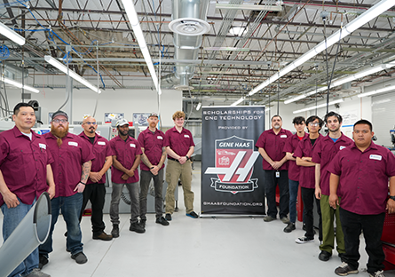 Group of 11 men in maroon work shirts standing to the left and right of a pull up banner with the Gene Haas logo