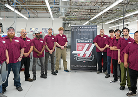 Group of 11 men in maroon work shirts standing to the left and right of a pull up banner with the Gene Haas logo