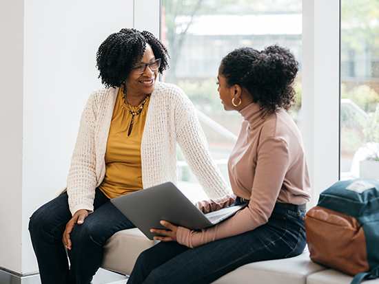 African American woman wearing a yellow top sitting next to younger woman holding a laptop mid conversation