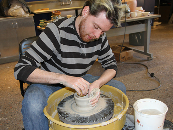 Photo of male student wearing black and grey striped long sleeve top. He is leaning over a yellow ceramics wheel forming clay with his hands. 