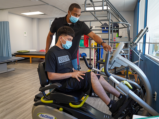 Photo of male physical therapist standing next to a male patient who is sitting on an exercise bike.