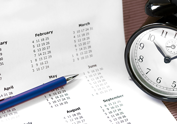 Monthly calendar on white paper with a blue pen and black analog clock sitting on a table.