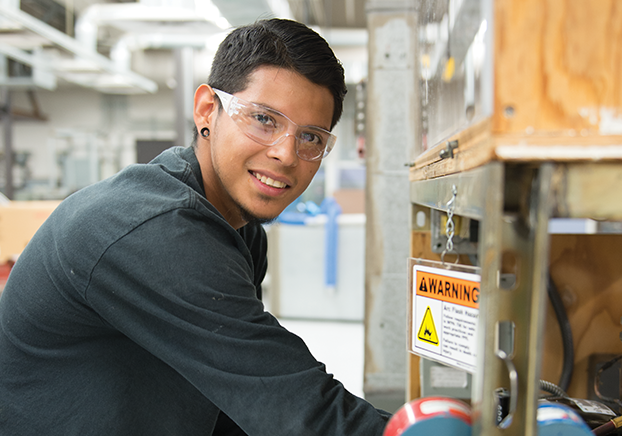 Male student kneeling down near an air compressor. He is wearing clear safety goggles.