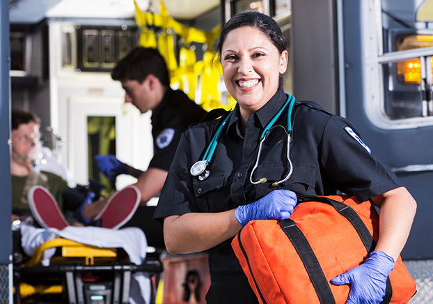 Female EMT carrying yellow duffle bag, wearing a stethoscope and blue gloves standing in front of an ambulance. 