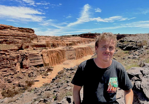 Photograph of young male wearing a black t-shirt sitting near the edge of a mountain with a canyon and mountain range behind him.